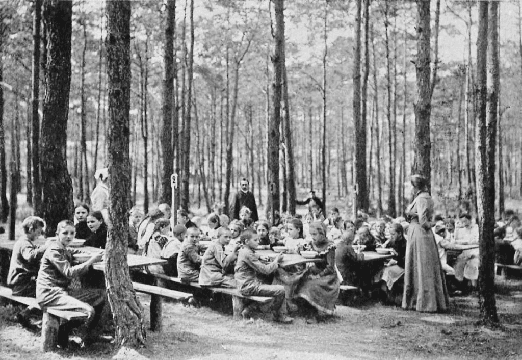 Black and white photo of students eating snack at large picnic tables at first ever open air school in Germany, 1904.